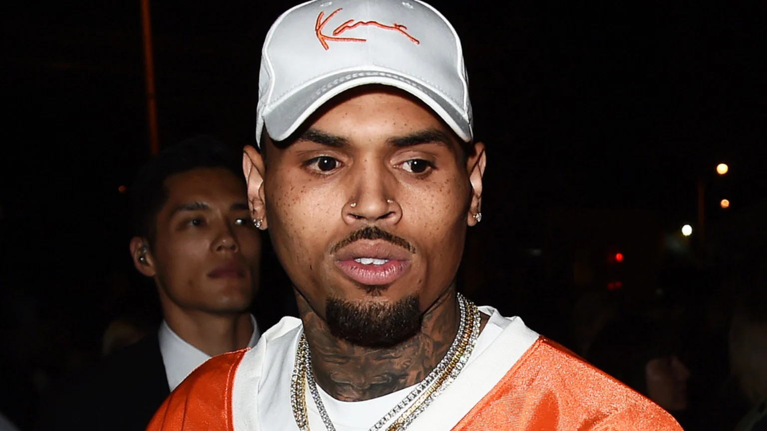 Chris Brown wears a white Kani cap, an orange and white shirt, and layered chains, looking forward; a man in a suit is partially visible behind him in a dark background with blurred lights.