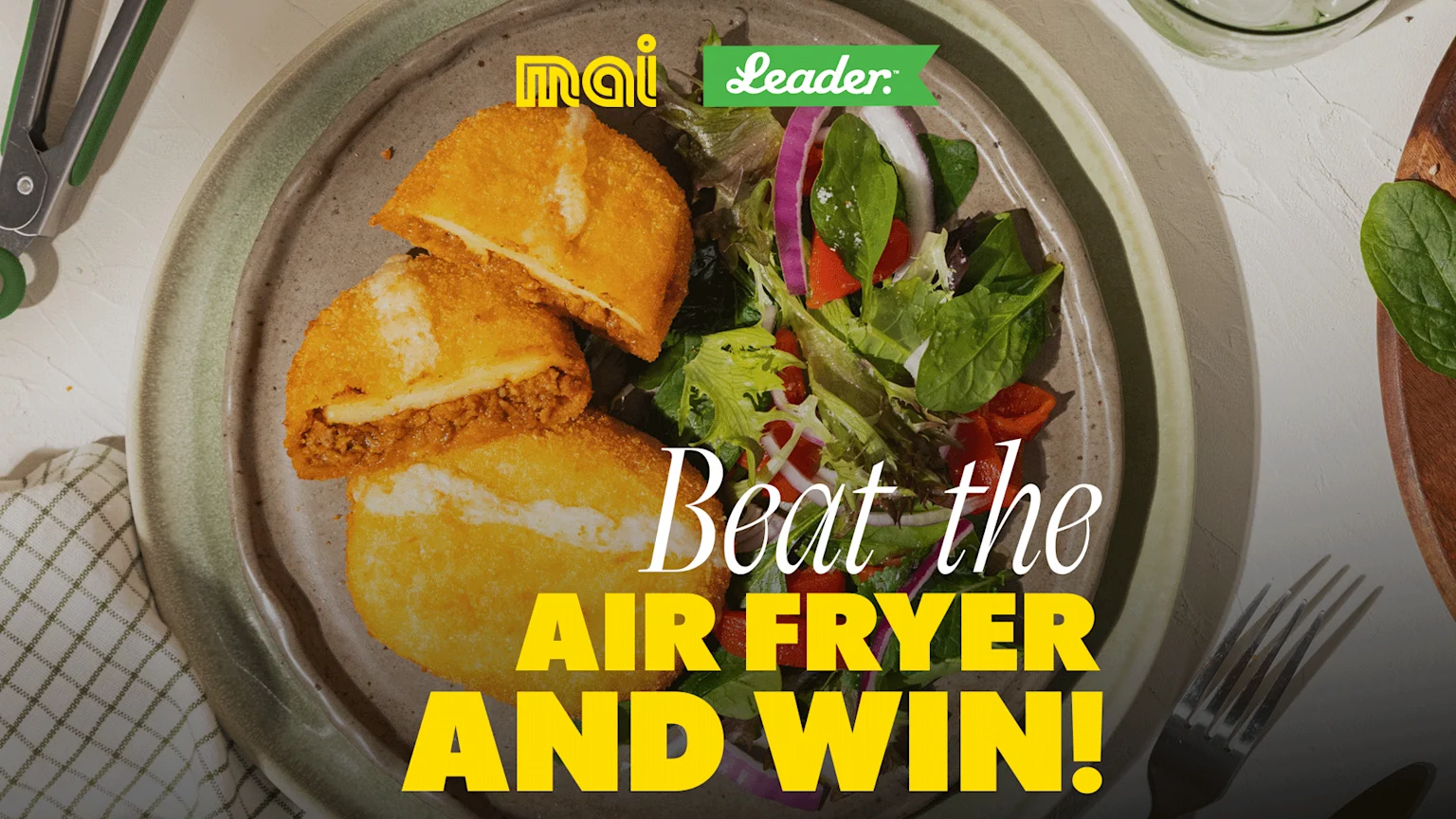 A plate holds two golden pastries, one cut open to show a meat filling, with a side salad, featuring "Beat the AIR FRYER AND WIN!" text and "mai" and "Leader" logos.