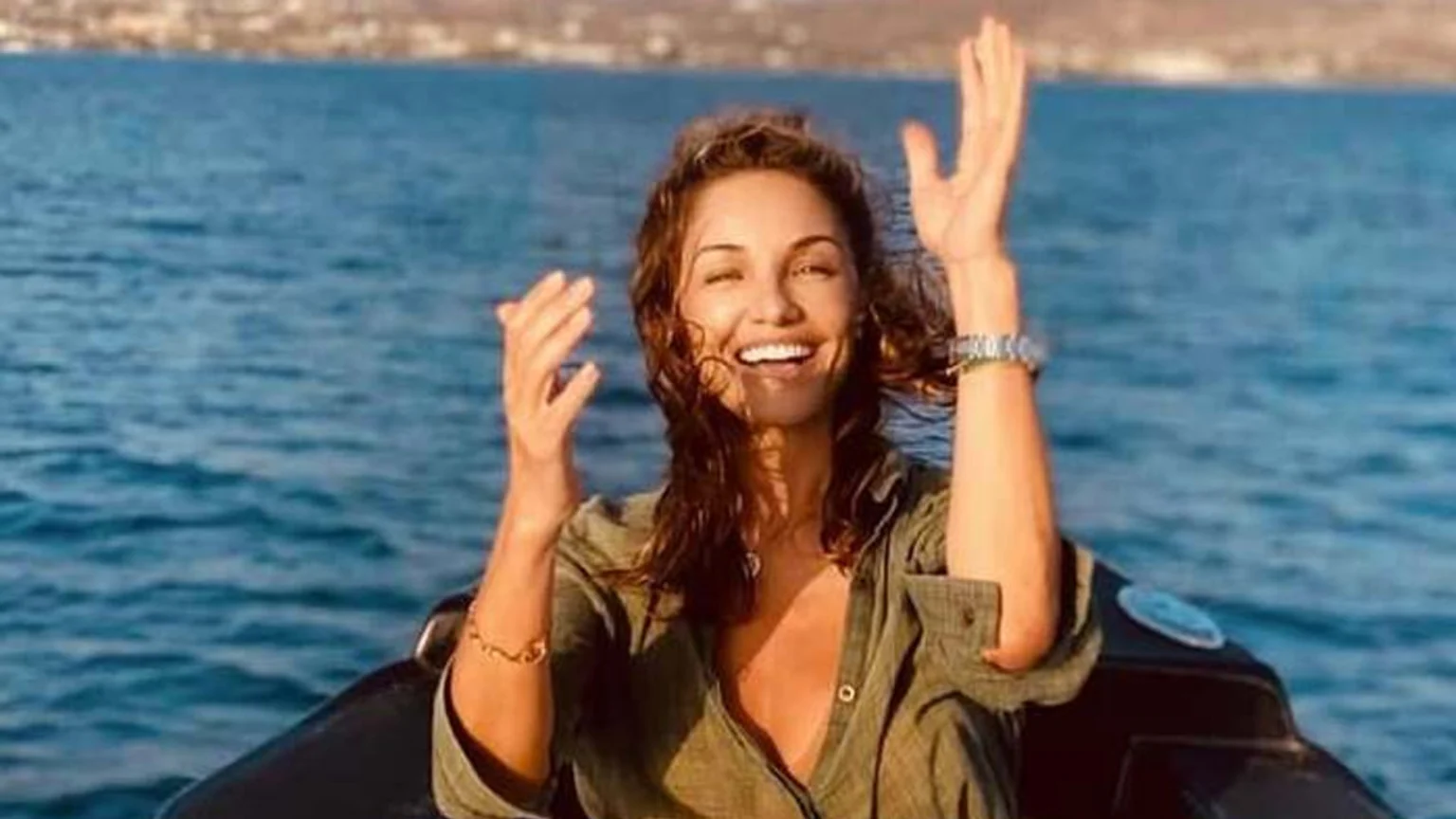 A smiling woman with wet brown hair, wearing a green shirt, raises her hands in a boat on blue water, with a coastline in the distance.