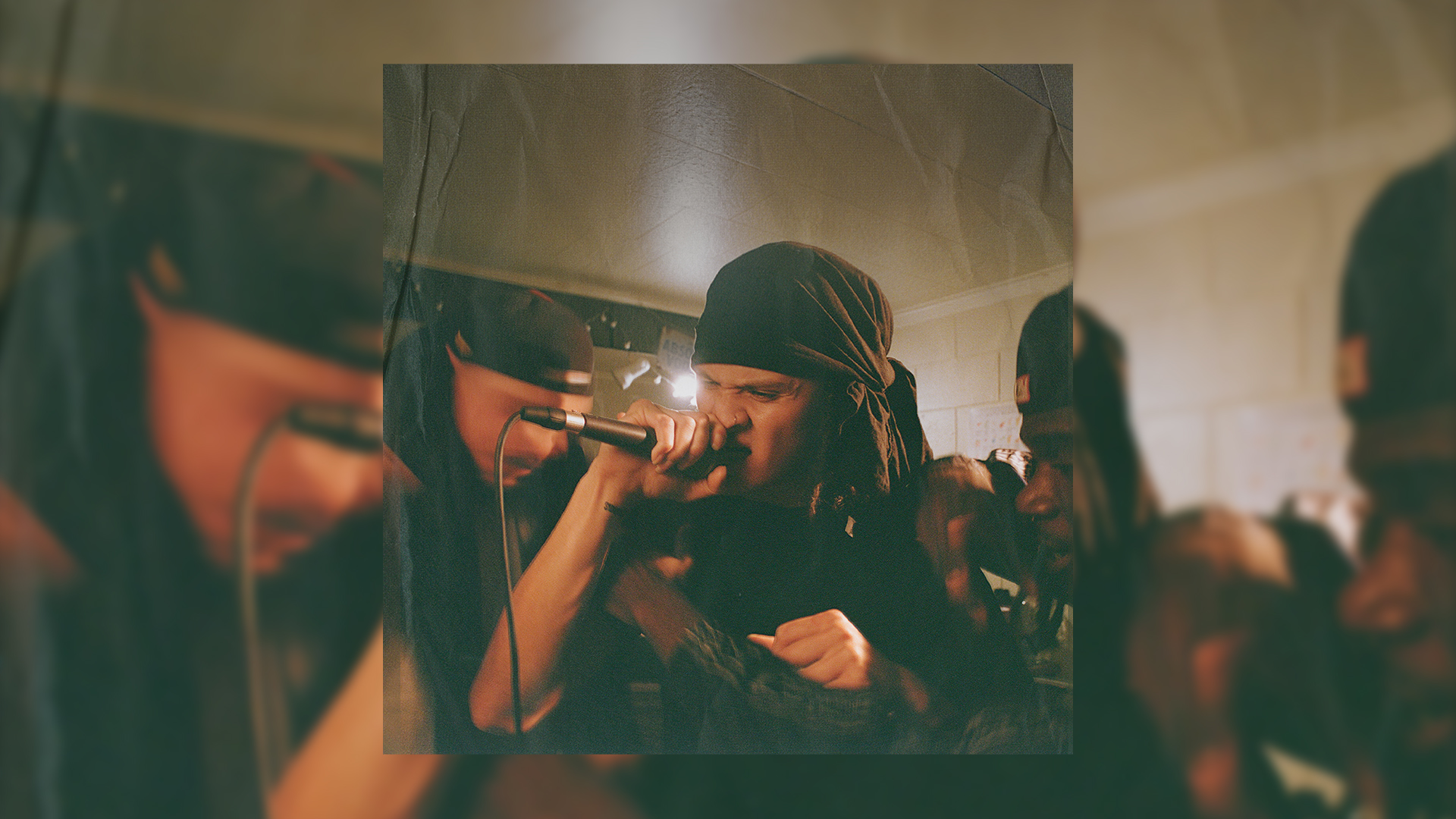 A performer with a dark head covering and dreadlocks sings intensely into a microphone in a grainy, low-light photo, with another person partially visible to the left.