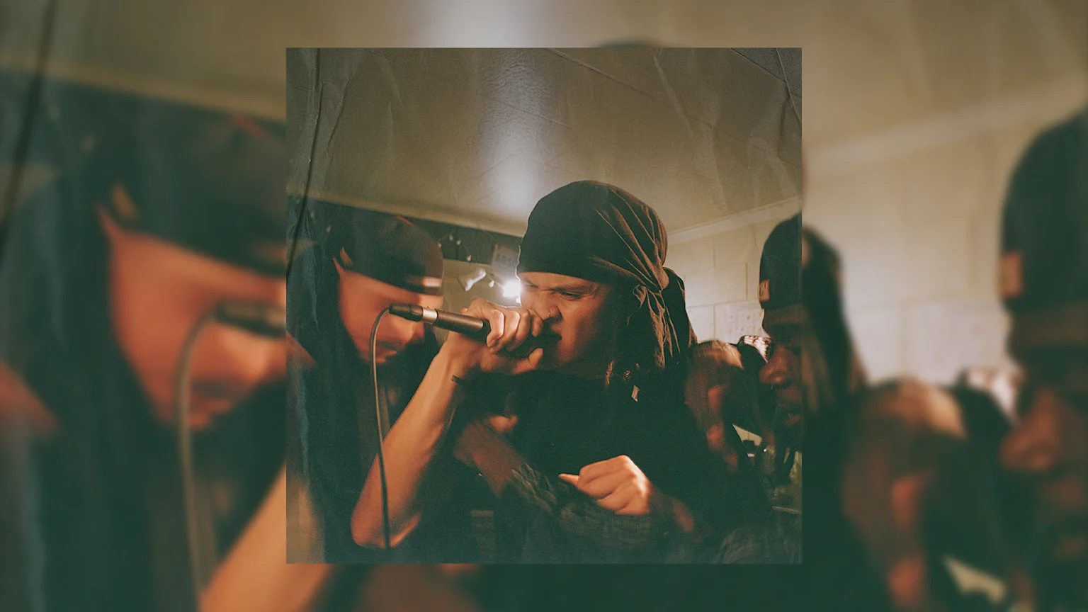 A performer with a dark head covering and dreadlocks sings intensely into a microphone in a grainy, low-light photo, with another person partially visible to the left.