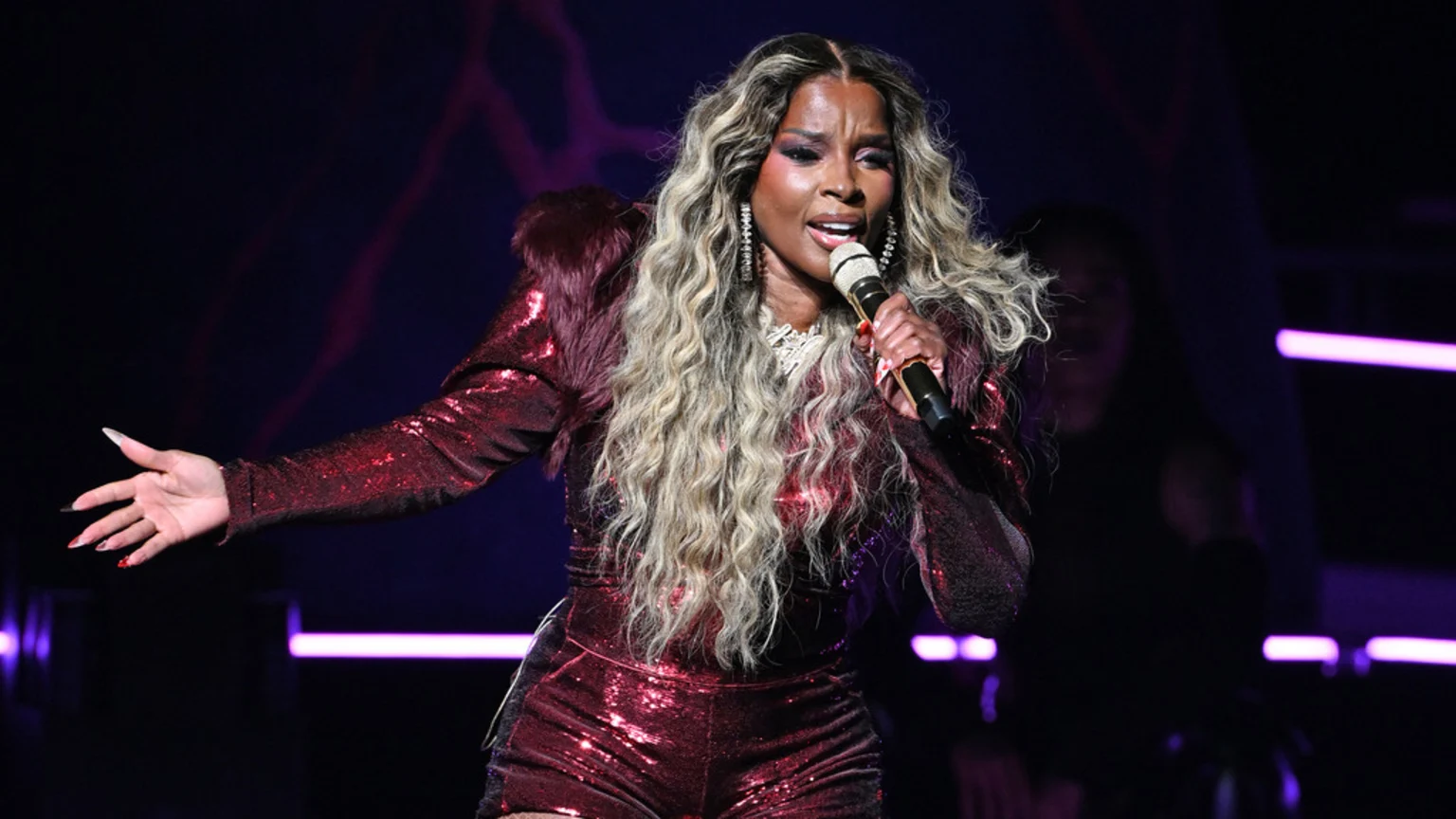 A singer with long wavy blonde hair and a shimmering dark red sequined outfit with fur shoulders performs on a dark stage, holding a microphone.