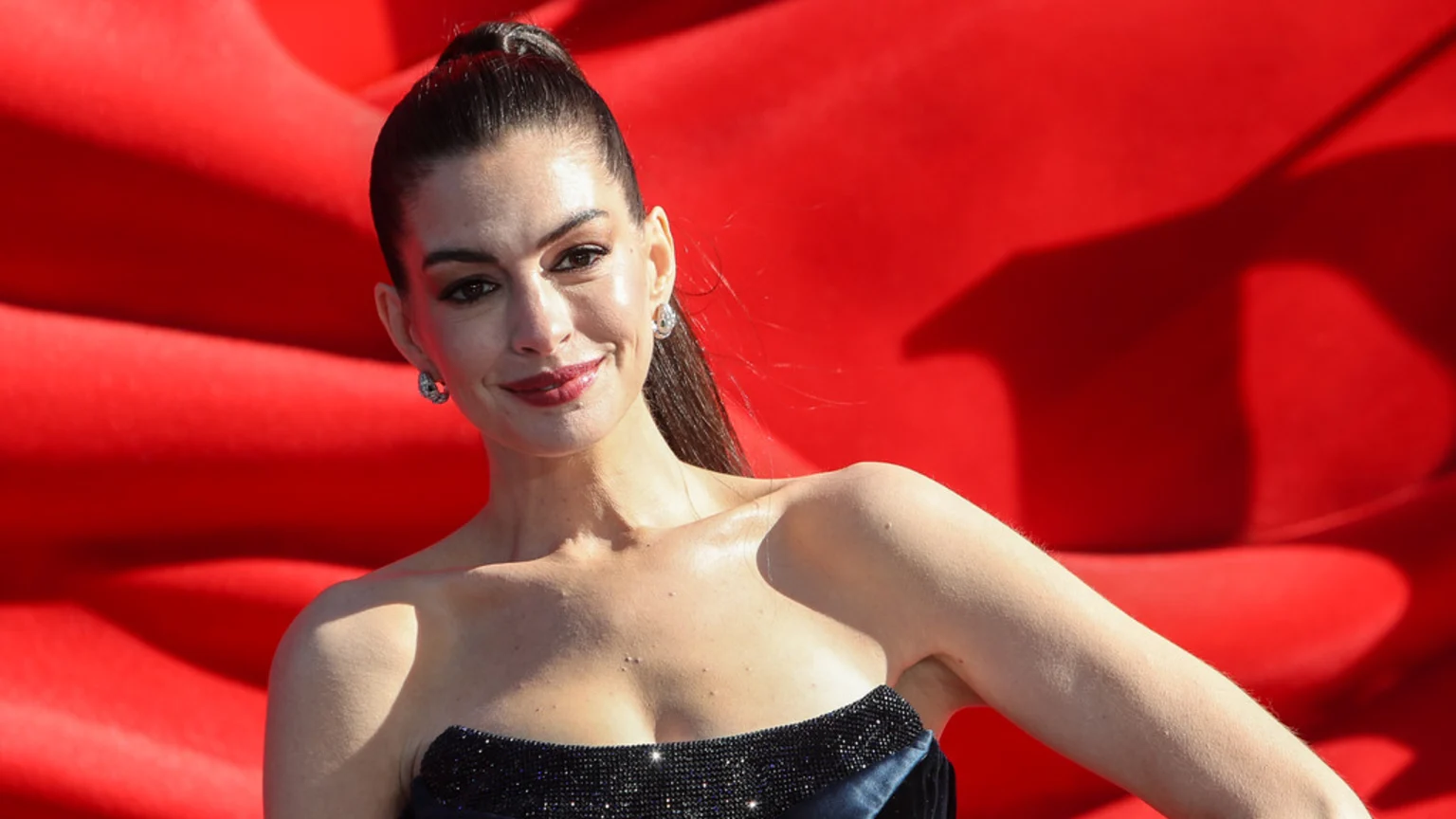 Anne Hathaway smiles at the camera, her dark hair in a high ponytail, wearing a dark, embellished strapless dress, against a vibrant red, textured background.