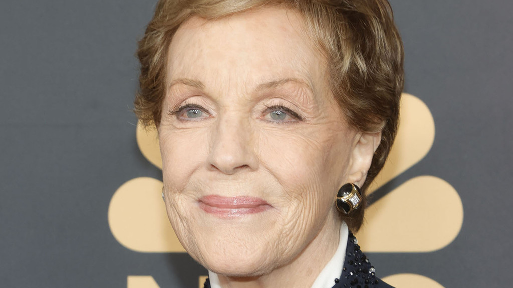 A close-up portrait of Julie Andrews with short blonde hair and blue eyes, smiling gently. She wears a dark top and a decorative earring against a grey background with gold shapes.