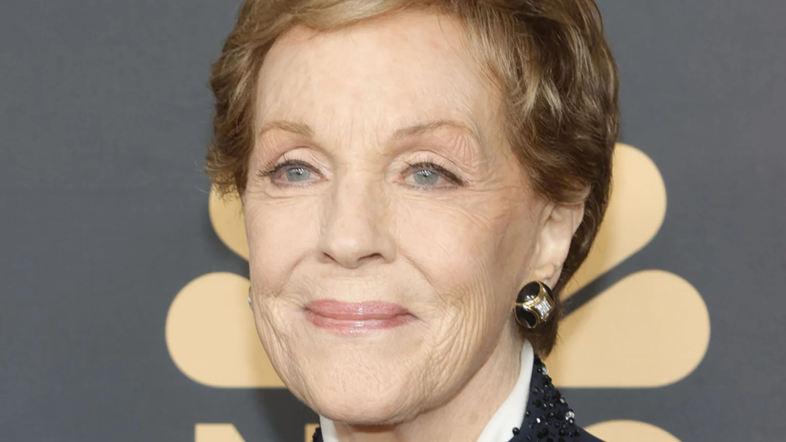 A close-up portrait of Julie Andrews with short blonde hair and blue eyes, smiling gently. She wears a dark top and a decorative earring against a grey background with gold shapes.