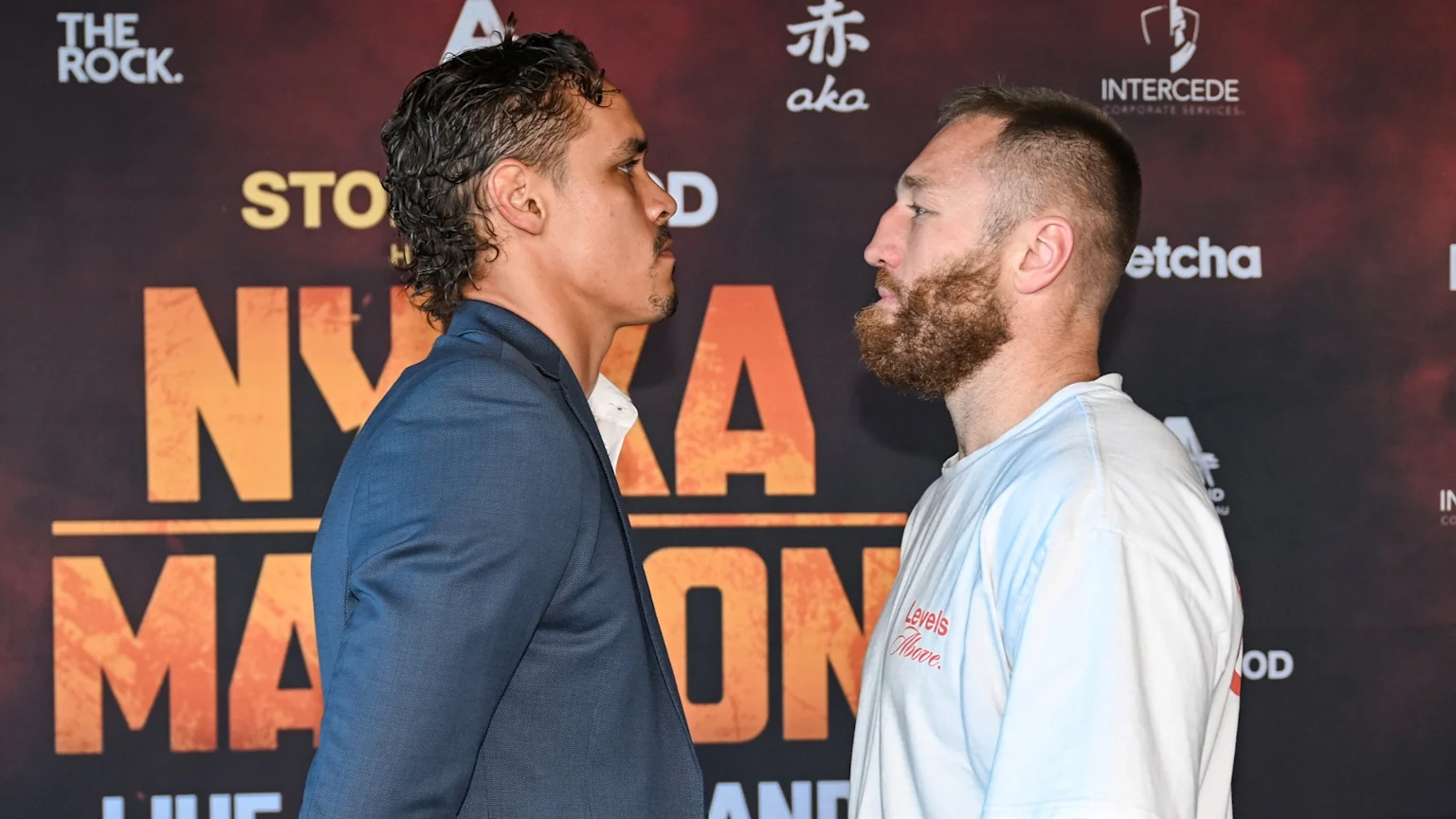 Two men in profile face each other intently, almost touching, against a dark background with orange lettering and sponsor logos; the man on the left wears a suit, the man on the right a t-shirt.
