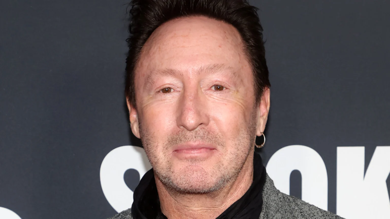 A close-up of Julian Lennon with dark hair, light brown eyes, and stubble, looking at the camera, wearing a dark top, grey jacket, and a small hoop earring.