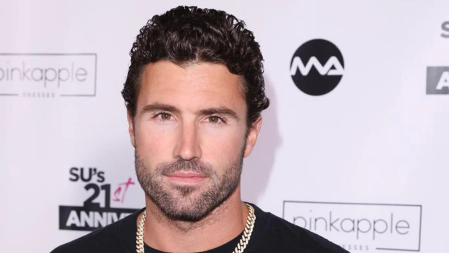 A man with dark curly hair and a beard looks forward, wearing a black shirt and a gold chain, against a white background with "pinkapple", "MA", and "SU's 21st ANNIV" logos.