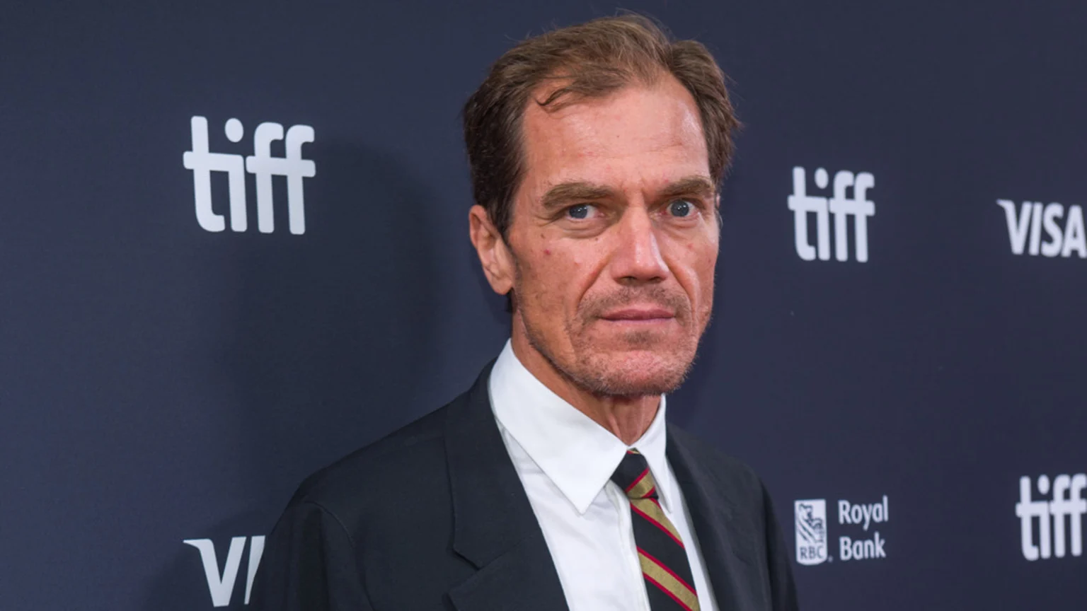 Actor Michael Shannon, wearing a dark suit, white shirt, and striped tie, looks directly at the camera against a dark blue background with white "tiff" and "VISA" logos.