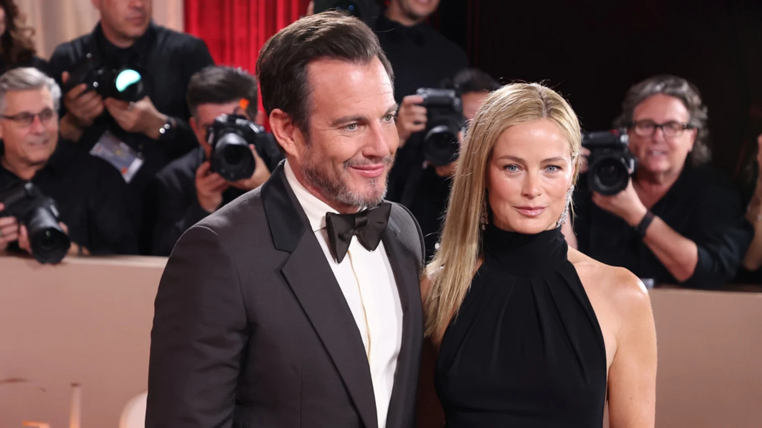 Will Arnett in a dark suit and bow tie smiles next to Carolyn Murphy in a black high-neck dress, both posing at a formal event with photographers in the background.