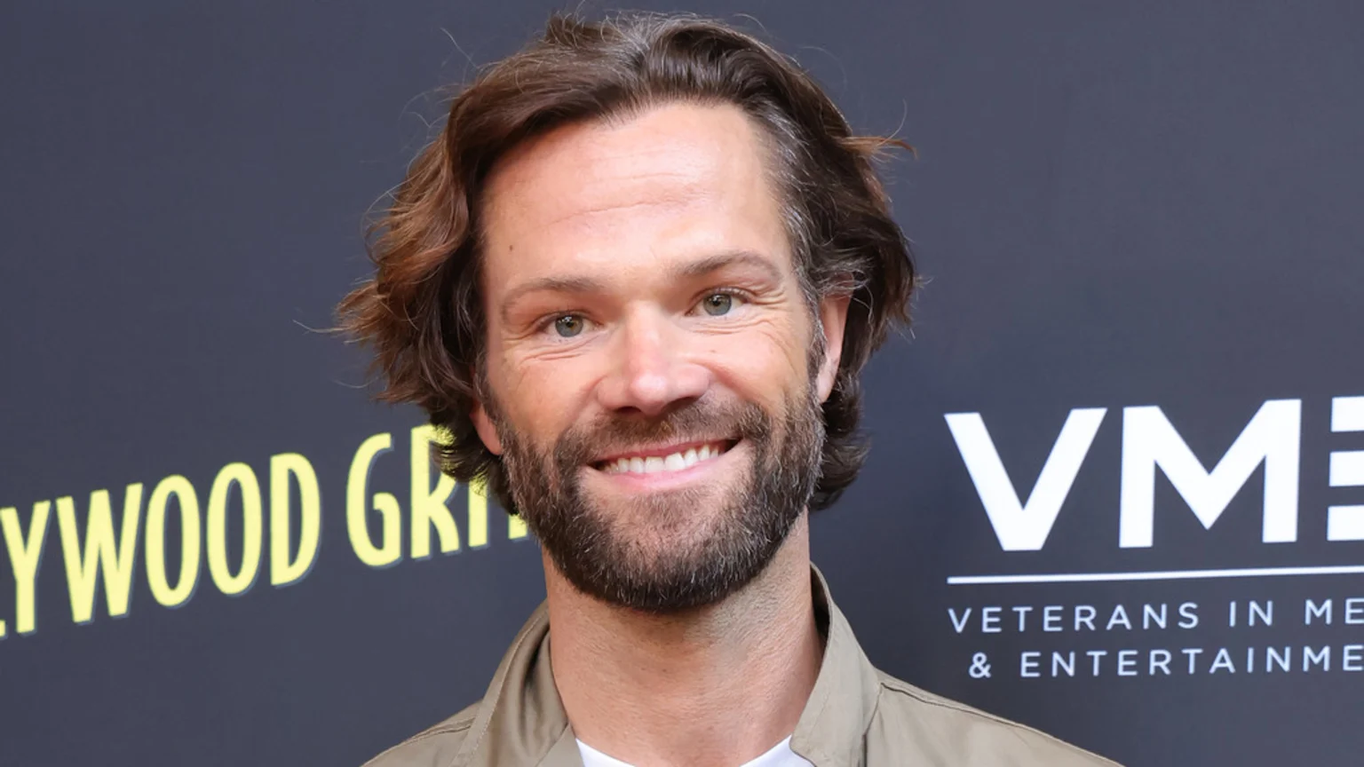Close-up of Jared Padalecki smiling, wearing a light brown jacket, with a dark background showing partial yellow and white text.