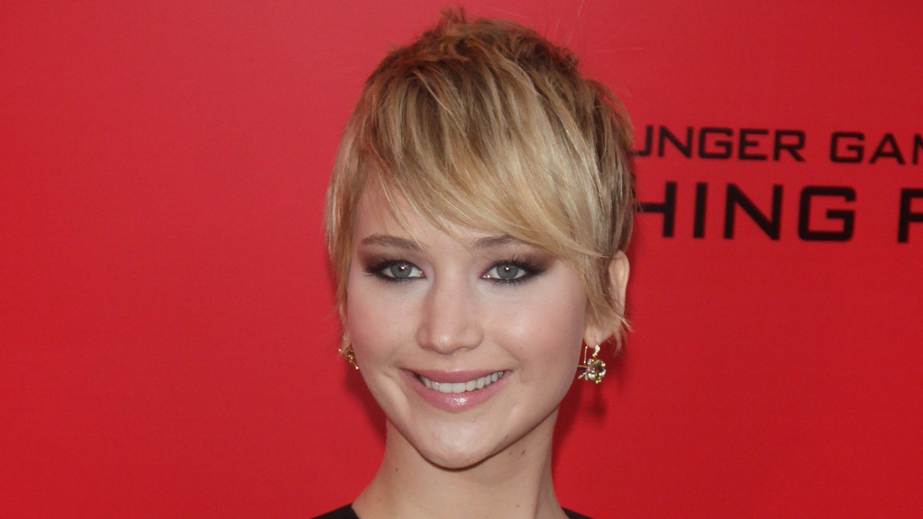 Close-up of Jennifer Lawrence with short, choppy blonde hair, dark eye makeup, and earrings, smiling against a red background with faint black text.