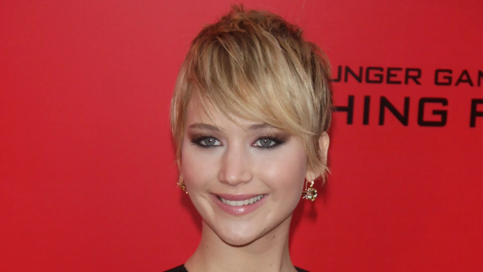 Close-up of Jennifer Lawrence with short, choppy blonde hair, dark eye makeup, and earrings, smiling against a red background with faint black text.