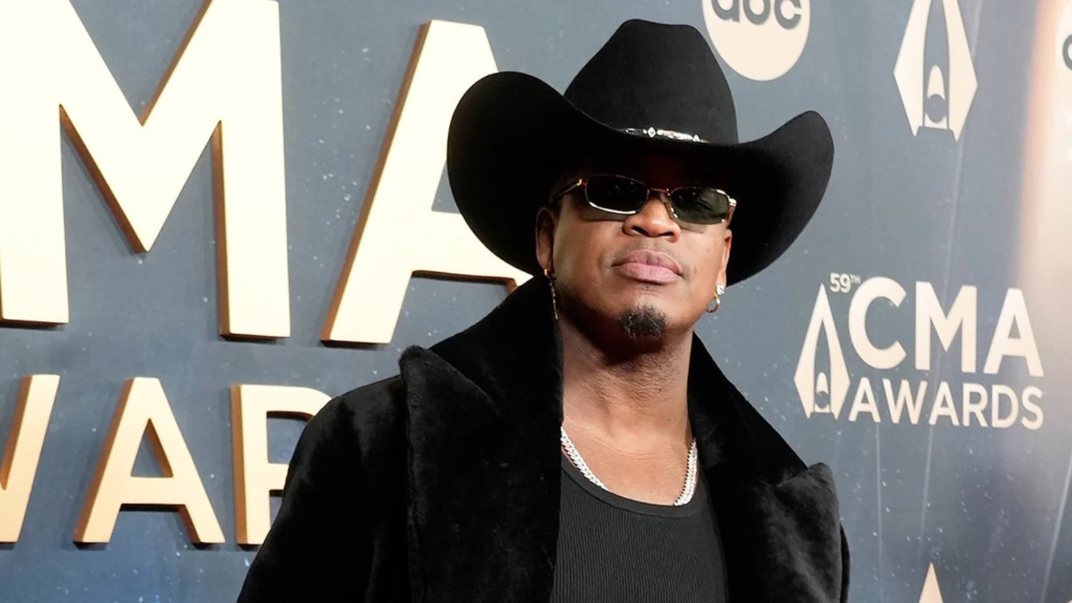 A man wearing a black cowboy hat, sunglasses, and a black faux fur coat poses in front of a backdrop displaying "CMA AWARDS" lettering.