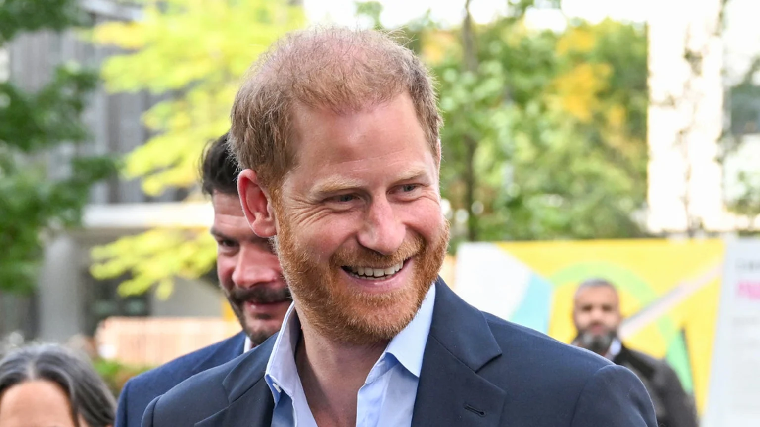 Prince Harry, a smiling man with red hair and a beard, wears a dark suit jacket and light blue shirt, with blurred people and green foliage in the background.