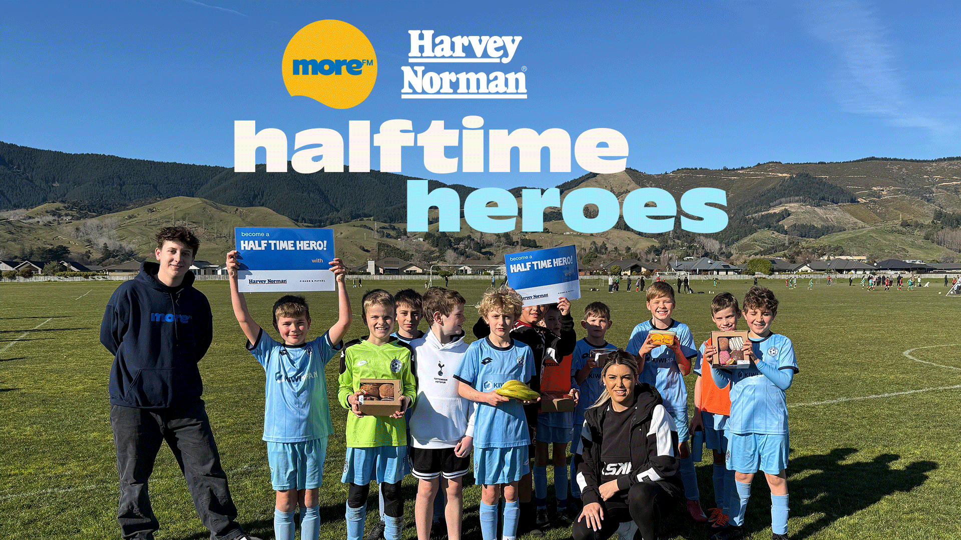 A group of boys and two adults pose on a sunny soccer field with mountains in the background, some boys hold "HALF TIME HERO!" signs and snacks, with "more™ Harvey Norman halftone heroes" text overlaid.