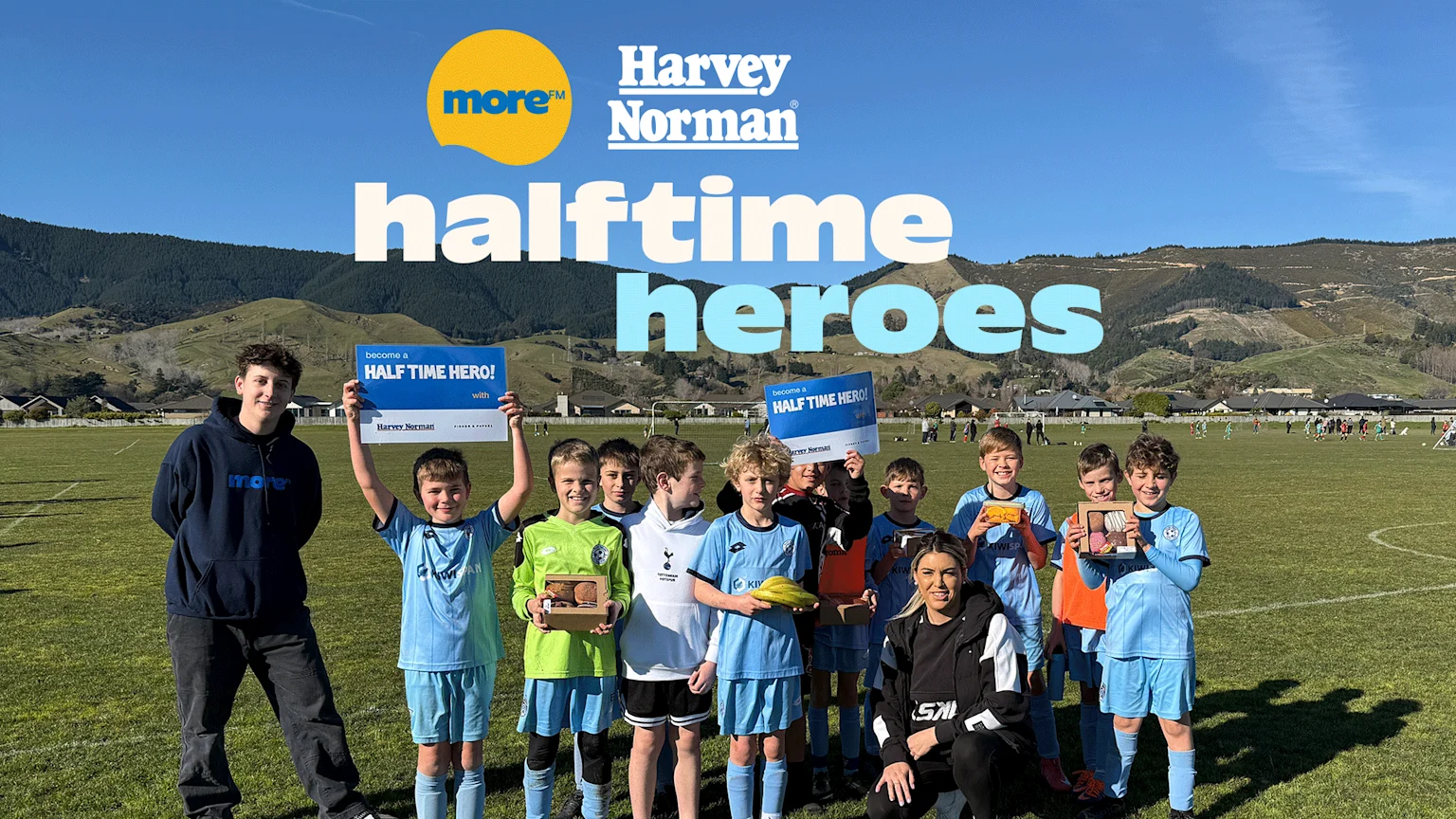 A group of boys and two adults pose on a sunny soccer field with mountains in the background, some boys hold "HALF TIME HERO!" signs and snacks, with "more™ Harvey Norman halftone heroes" text overlaid.