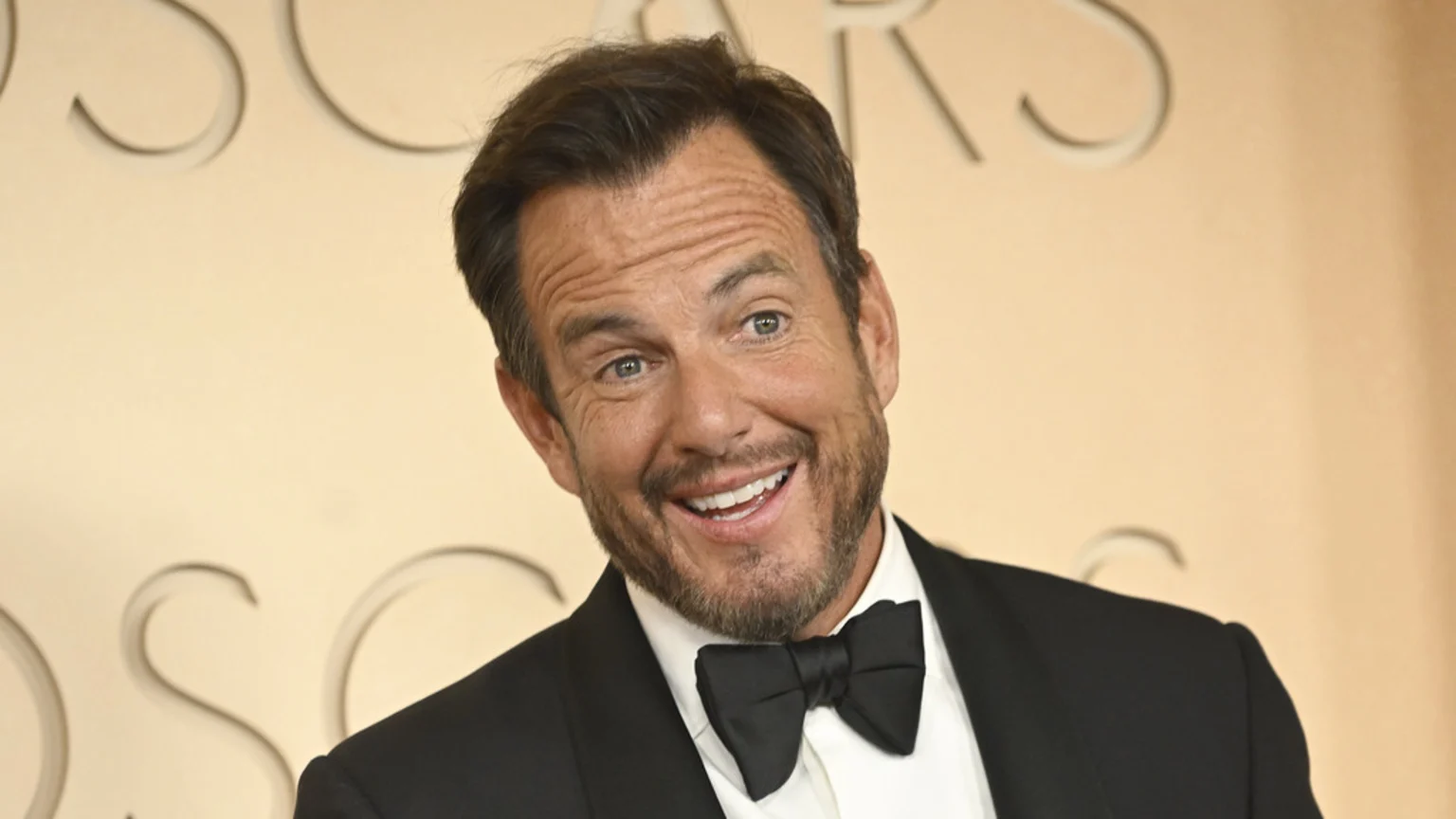 Will Arnett smiles broadly, showing his teeth, while wearing a black tuxedo and bow tie against a light background with faint embossed letters.