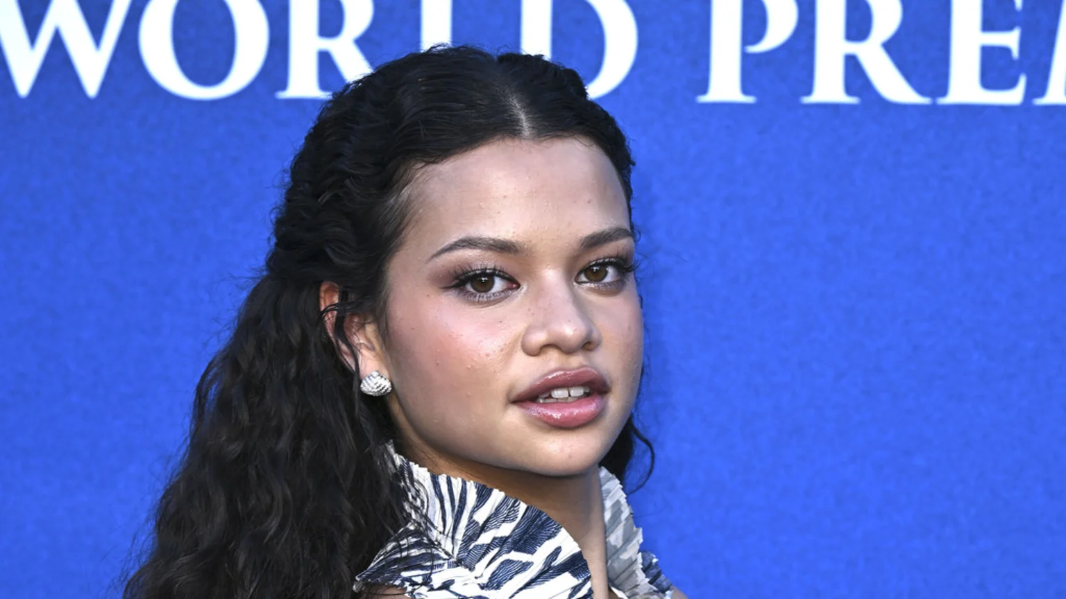 Catherine Laga'aia, a young woman with dark curly hair, light makeup, and a patterned top, looks slightly right against a blue "WORLD PREMIERE" banner.