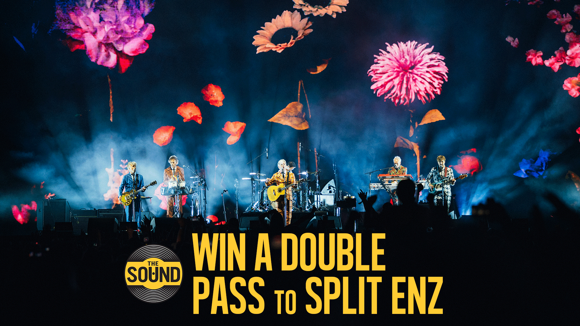 A band performs on a stage illuminated by smoke and colorful flower projections, with an audience in the foreground and text overlay "WIN A DOUBLE PASS TO SPLIT ENZ".
