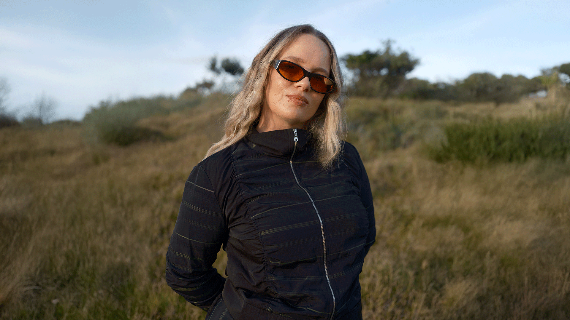 A blonde woman in orange sunglasses and a dark zip-up jacket stands in a grassy field under a cloudy sky, looking at the camera.