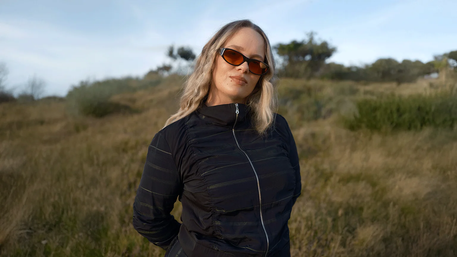 A blonde woman in orange sunglasses and a dark zip-up jacket stands in a grassy field under a cloudy sky, looking at the camera.