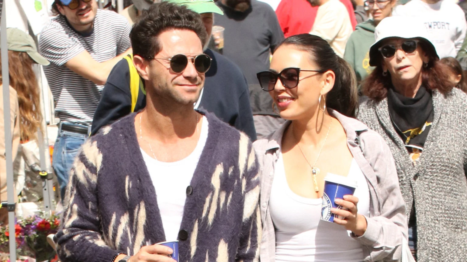 Janel Parrish and Sasha Farber, a woman in sunglasses holding a coffee cup and a man in sunglasses and a patterned cardigan, smile while walking outdoors.