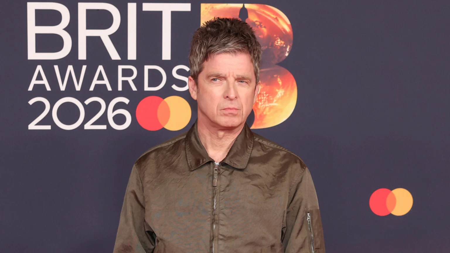 Noel Gallagher, a man with grey hair, stands in a dark olive jacket against a dark background featuring "BRIT AWARDS 2026" text and the Mastercard logo.