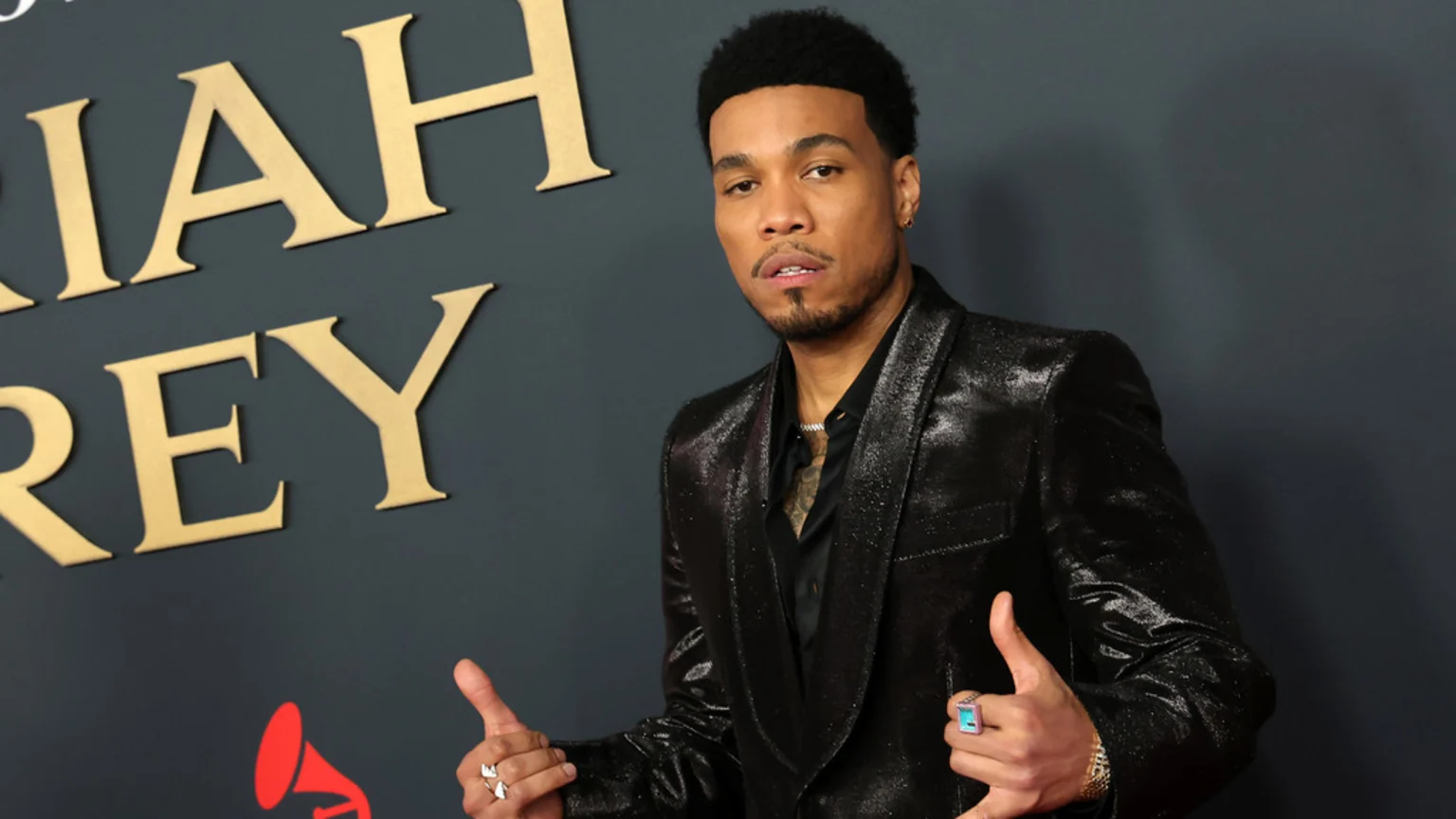 A man in a shiny black suit with tattoos and rings smiles with thumbs up, standing in front of a dark grey wall with gold letters spelling "MARIAH CAREY" and a red gramophone logo.