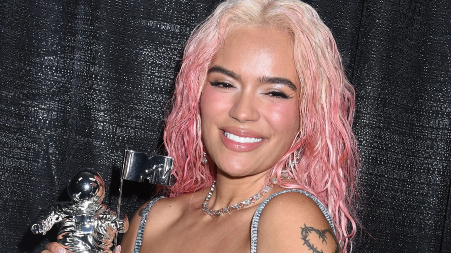 Karol G smiles, holding a silver MTV VMA Moon Person award, showing her pink and blonde wavy hair, sparkly top, and a barbed wire heart tattoo on her arm.