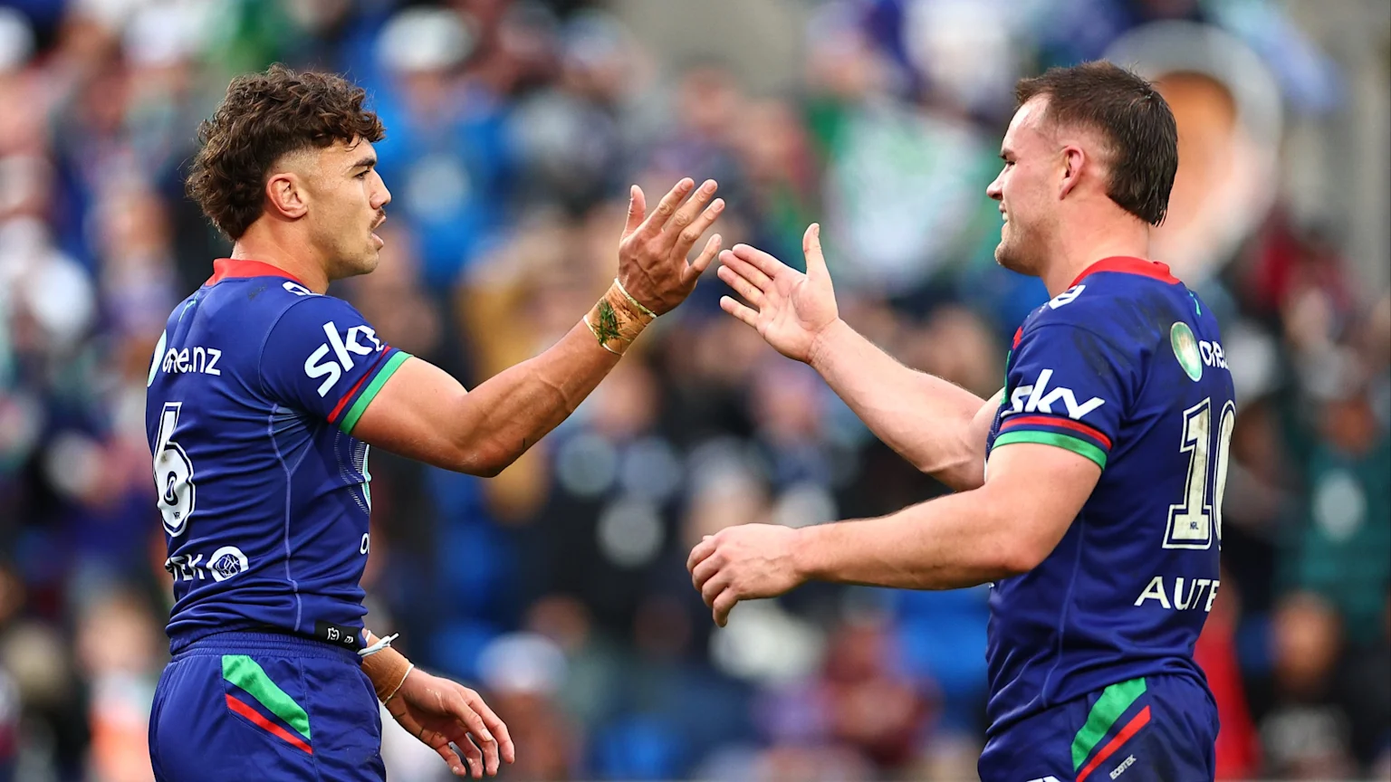 NRL Warriors player Chanel Harris-Tavita (number 6) with curly hair high-fives a teammate (number 10) in blue jerseys on a blurred background.