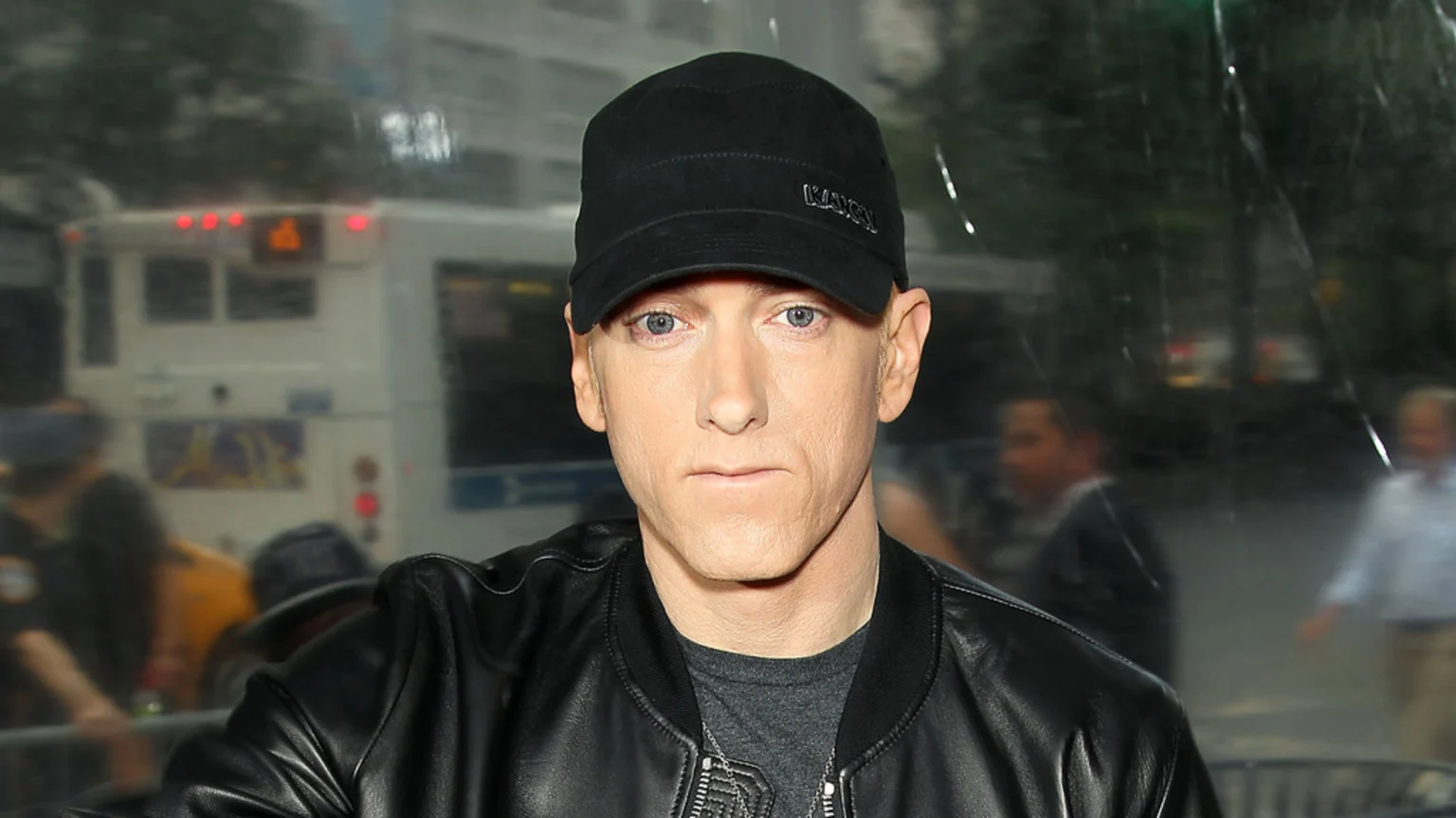Eminem wears a black KANGOL cap and a black leather jacket, looking straight ahead with blue eyes, against a blurred urban background with a bus.