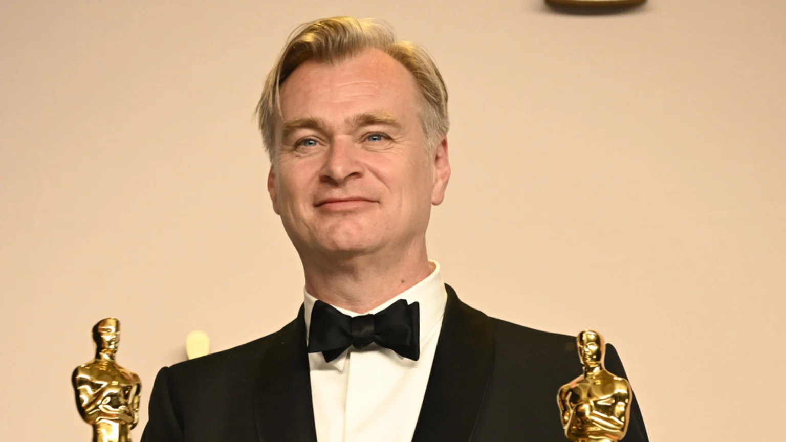 Christopher Nolan, smiling in a tuxedo with a bow tie, holds two golden Oscar statuettes, one on each side, against a plain background.