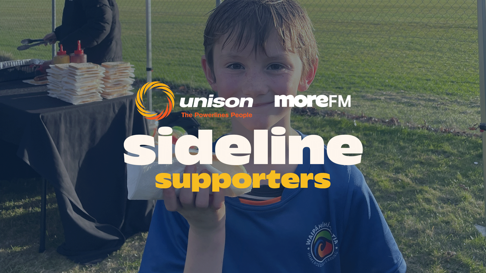 A smiling boy holds a hotdog in front of a BBQ table with food, buns, and sauces, with "sideline supporters" text and sponsor logos visible.
