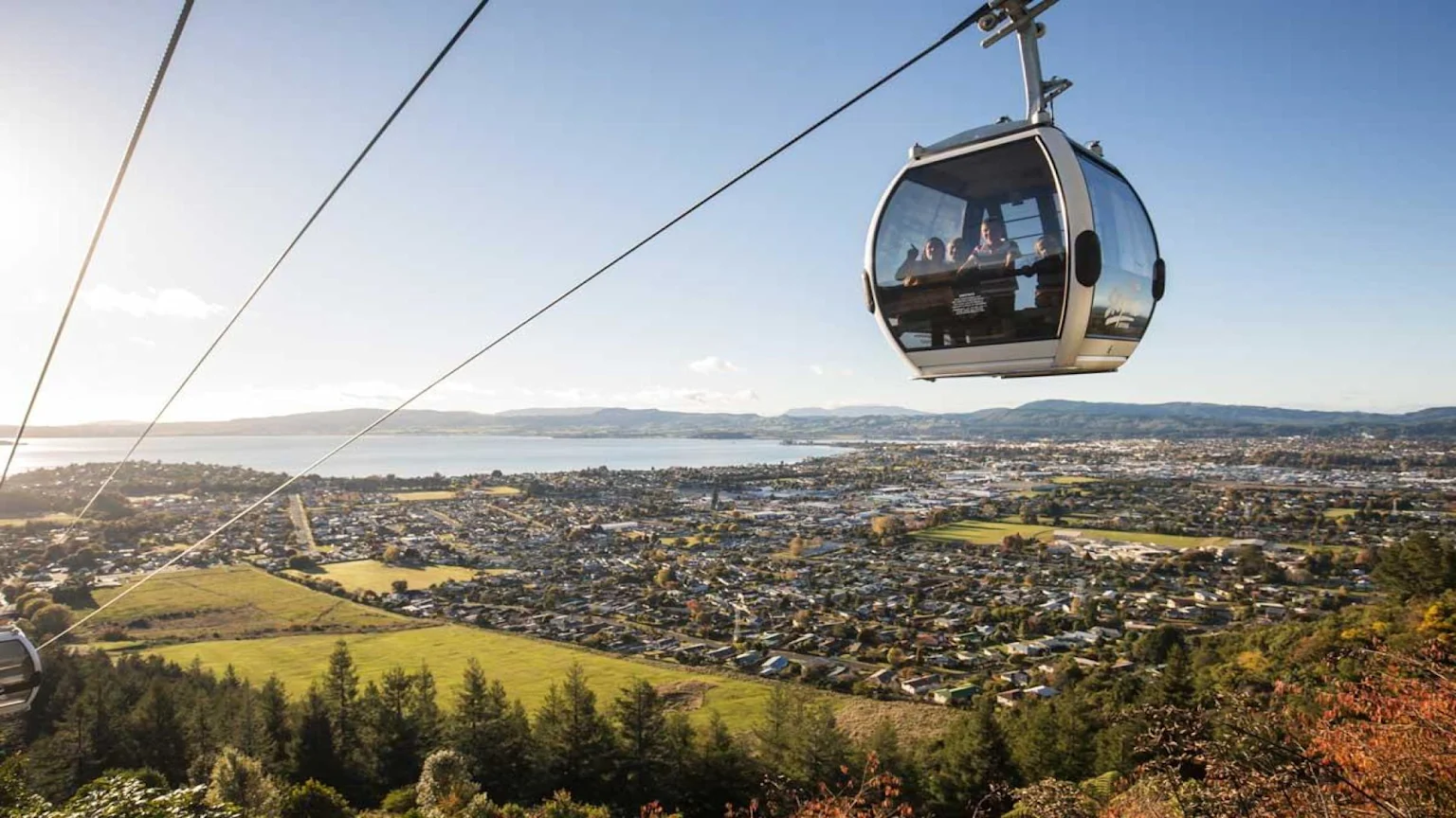 A gondola cabin carries passengers high above a sprawling city and a large lake backed by mountains, under a bright blue sky, offering a vast panoramic view.