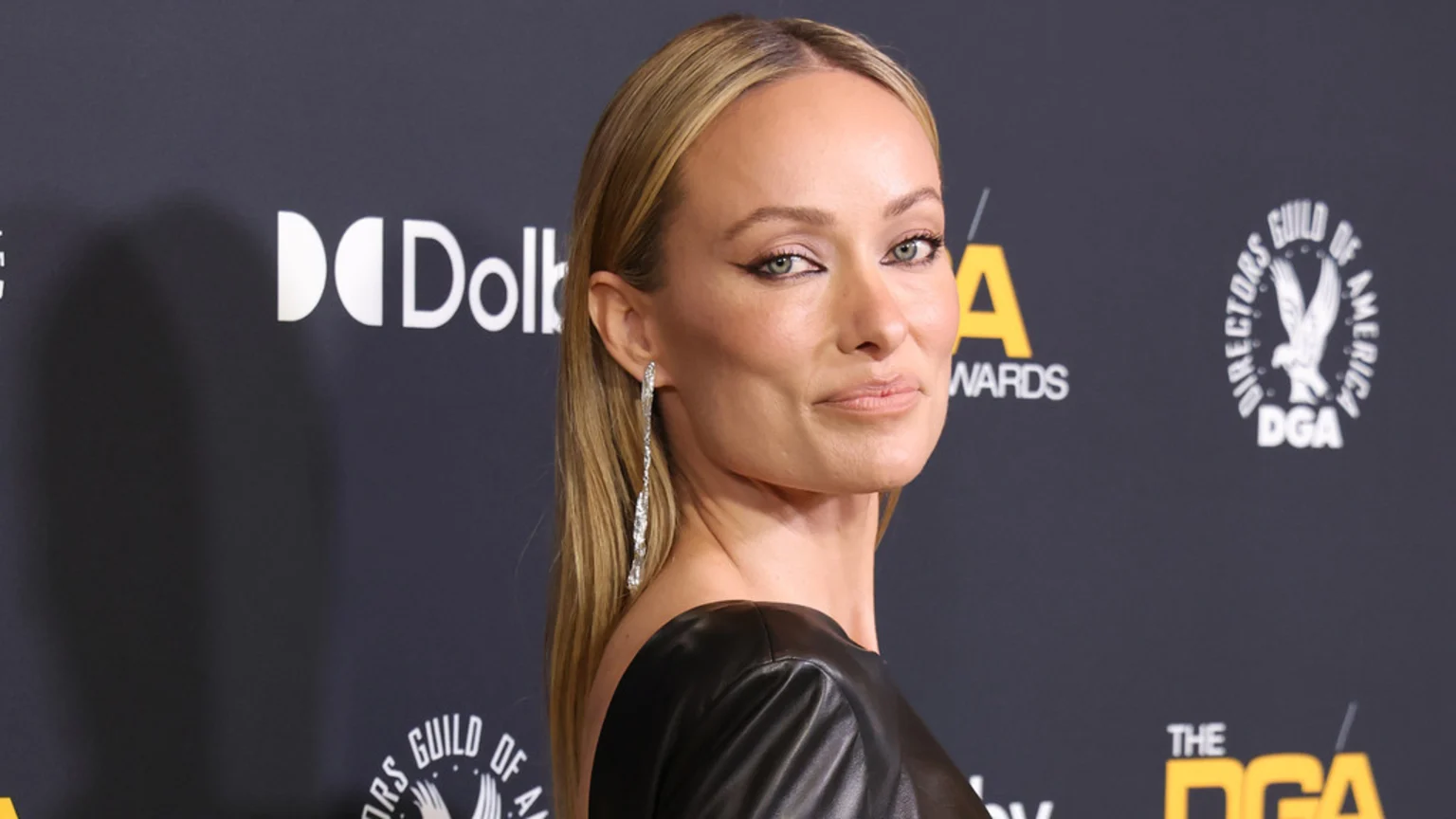Olivia Wilde, a blonde woman, looks towards the viewer, wearing dramatic winged eyeliner, long diamond earrings, and a dark top, with "Dolby" and "DGA" logos in the background.