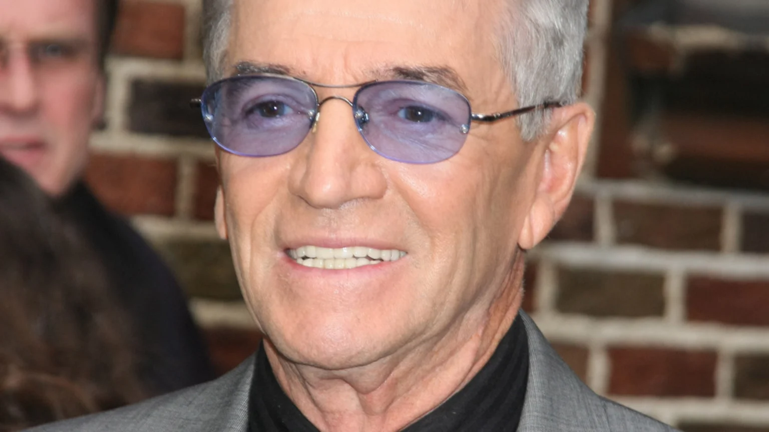 A close-up portrait of an older man with gray hair and purple-tinted sunglasses, smiling and showing his teeth, wearing a gray suit and a dark shirt, with a blurred person and a brick wall in the background.