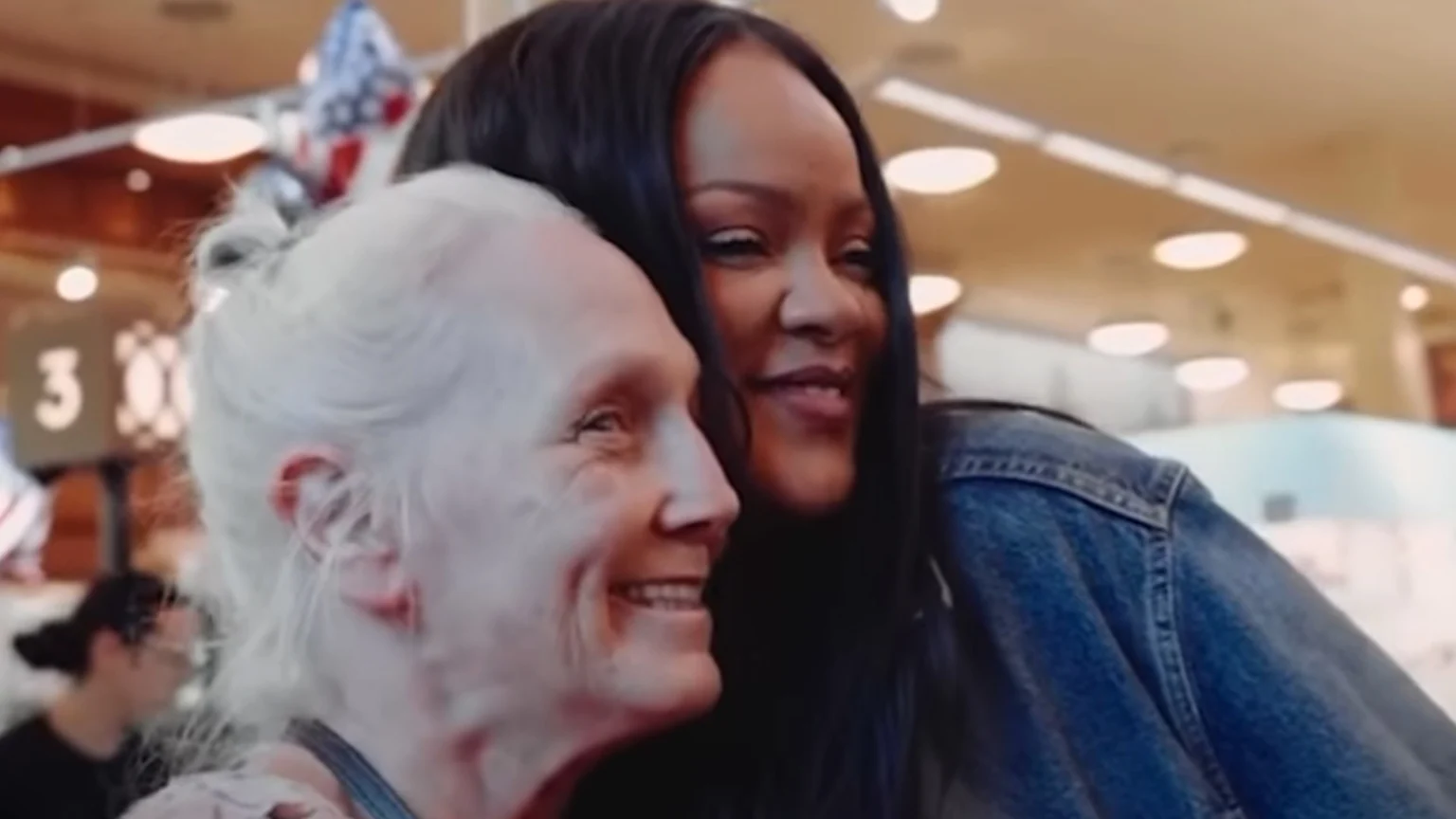 Two women, one with long dark hair and a denim jacket and the other with short white hair, embrace and smile warmly in an indoor setting.