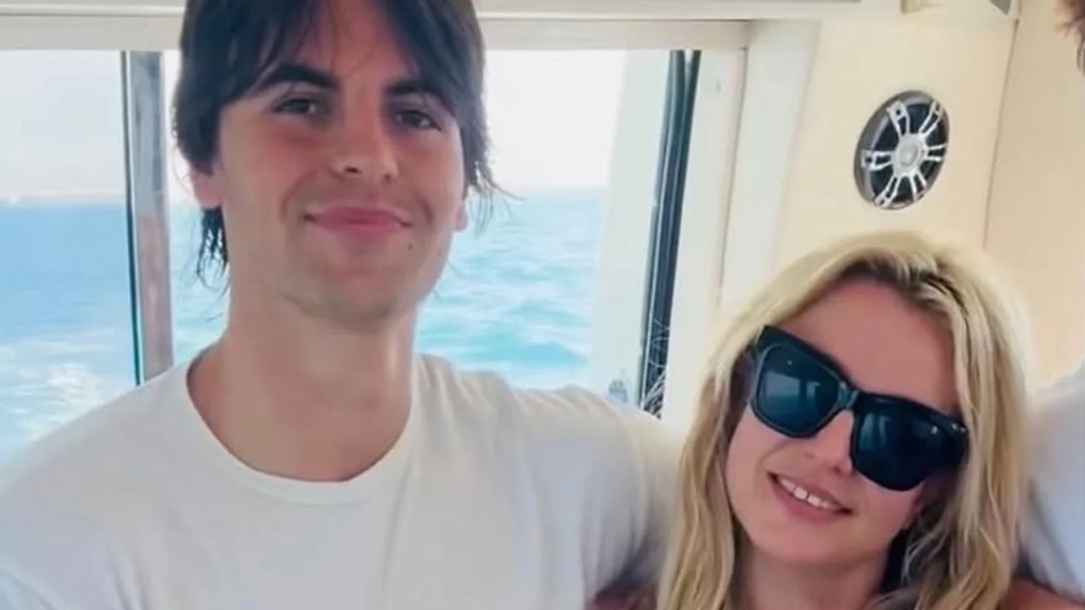 A smiling young man with dark hair and a blonde woman wearing dark sunglasses pose for a photo on a boat with the ocean visible behind them.