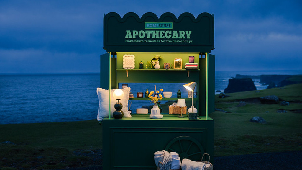 A green "HomeSense Apothecary" display cart filled with cozy homeware like lamps, candles, and blankets, stands illuminated by the dark ocean and cliffs at dusk.