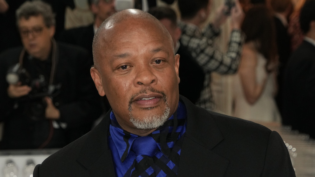 A man with dark skin, a shaved head, and a gray goatee, identified as Dr. Dre, wears a black suit and a blue patterned shirt with a tie, looking slightly to the right.