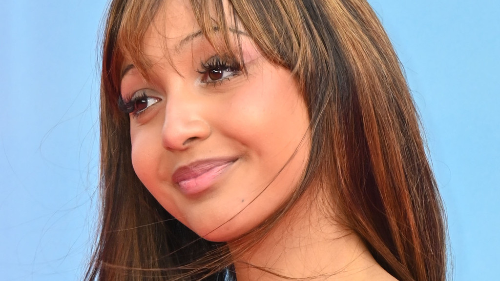 A close-up portrait of an unknown person with medium-brown skin, long brown hair with bangs, and makeup, gently smiling against a plain light blue background. The person is wearing a red/orange top and looking slightly upwards and left.