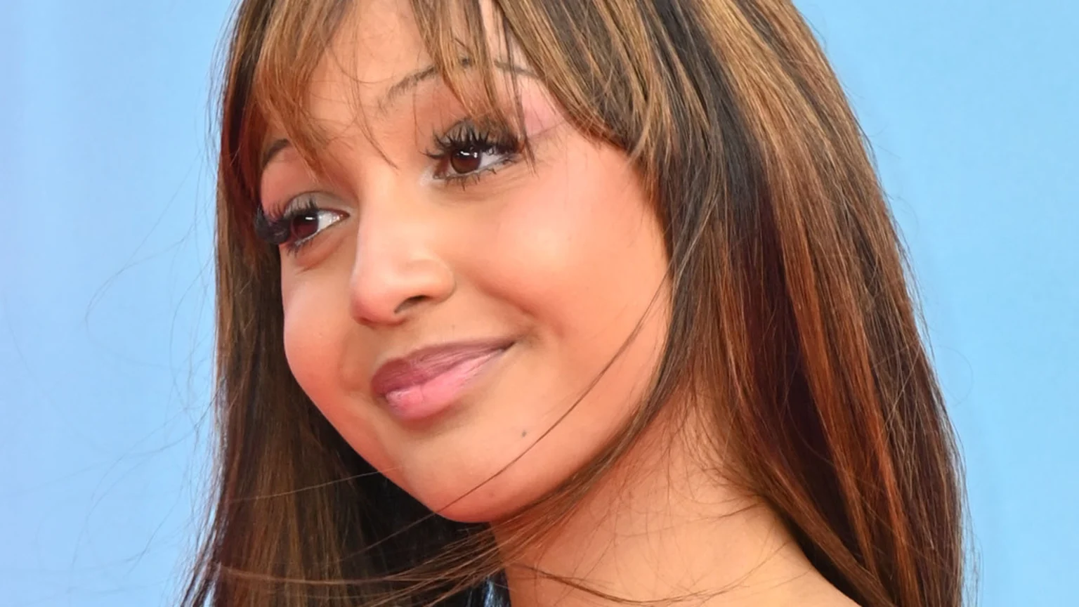 A close-up portrait of an unknown person with medium-brown skin, long brown hair with bangs, and makeup, gently smiling against a plain light blue background. The person is wearing a red/orange top and looking slightly upwards and left.