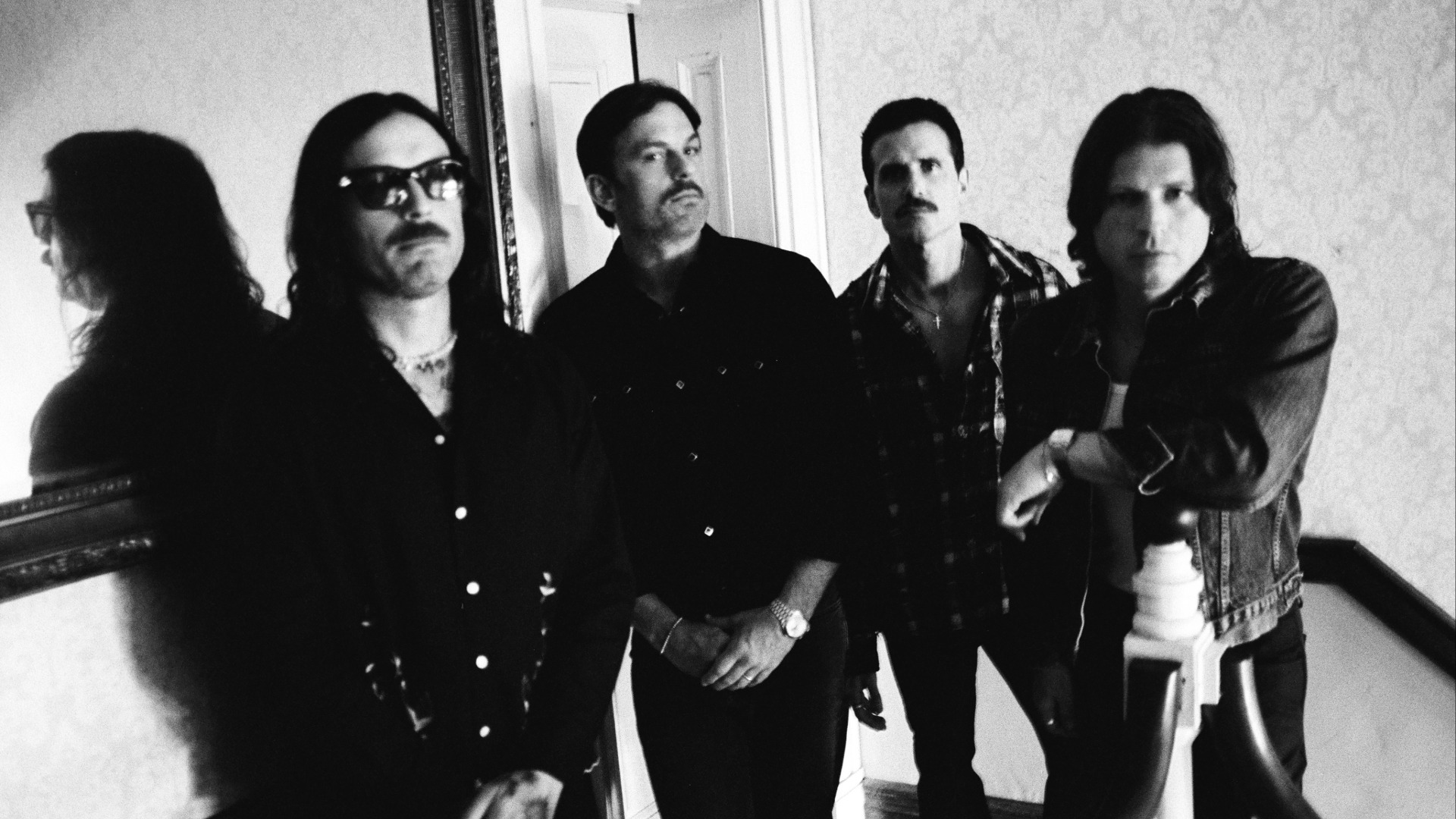 Black and white photo of four men posing indoors. The first wears sunglasses and a mustache, the second and third have mustaches, and the fourth leans on a banister.