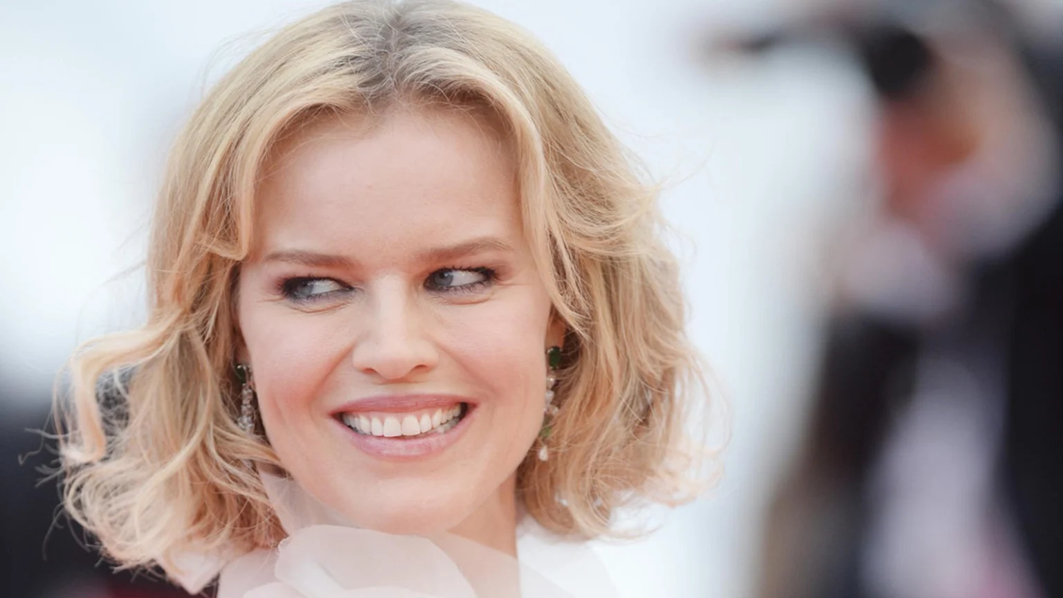 A smiling blonde woman with wavy hair and blue eyes looks to the side, wearing pearl and green stone drop earrings and a light-colored top.