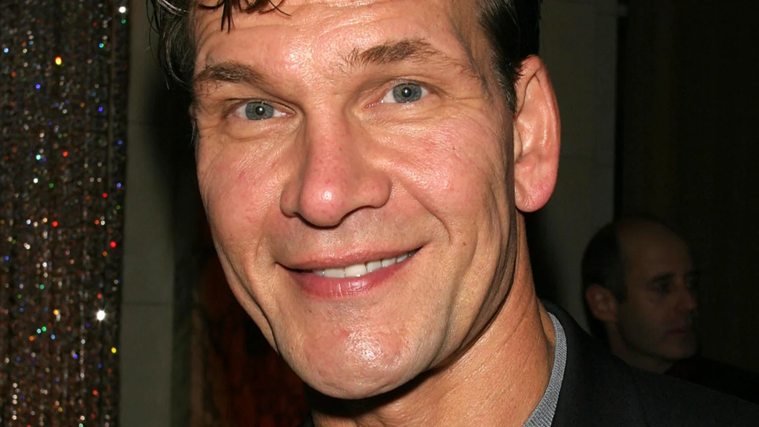 Close-up headshot of actor Patrick Swayze smiling directly at the camera, with light blue eyes and dark hair, against a blurry background with colorful lights.