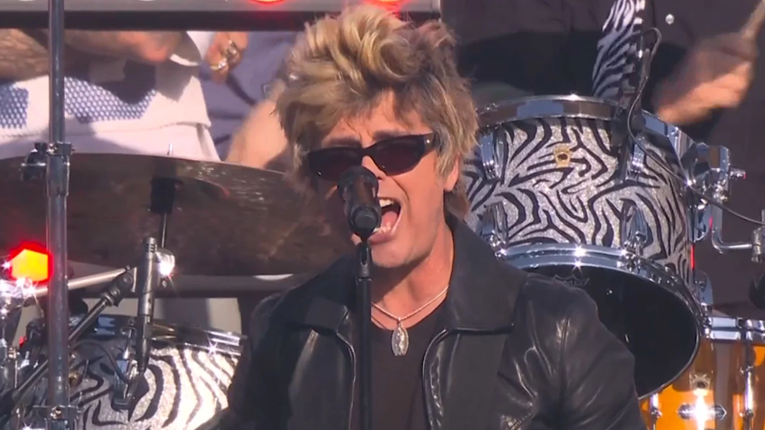 A male vocalist with spiky blonde hair and dark sunglasses sings into a microphone, wearing a black leather jacket and silver necklace. The background shows a drummer playing a zebra-print drum set with visible REMO branding under bright stage lighting.