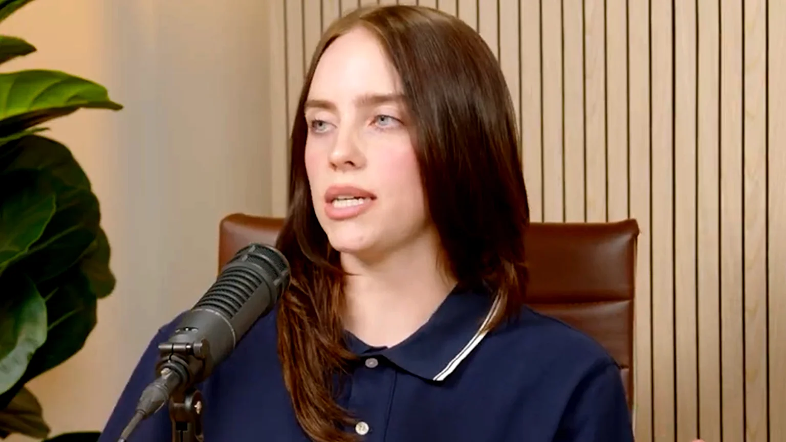 Billie Eilish, with long dark hair, wears a blue shirt, sitting behind a desk and microphone.