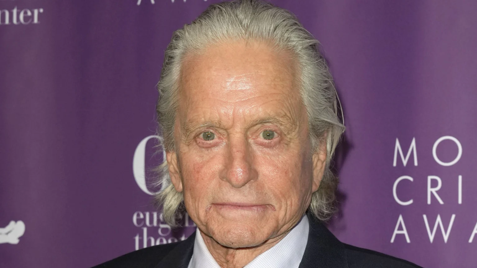 A close-up portrait of actor Michael Douglas with grey hair and green eyes, looking directly at the camera with a neutral expression, wearing a dark suit jacket over a white collared shirt.