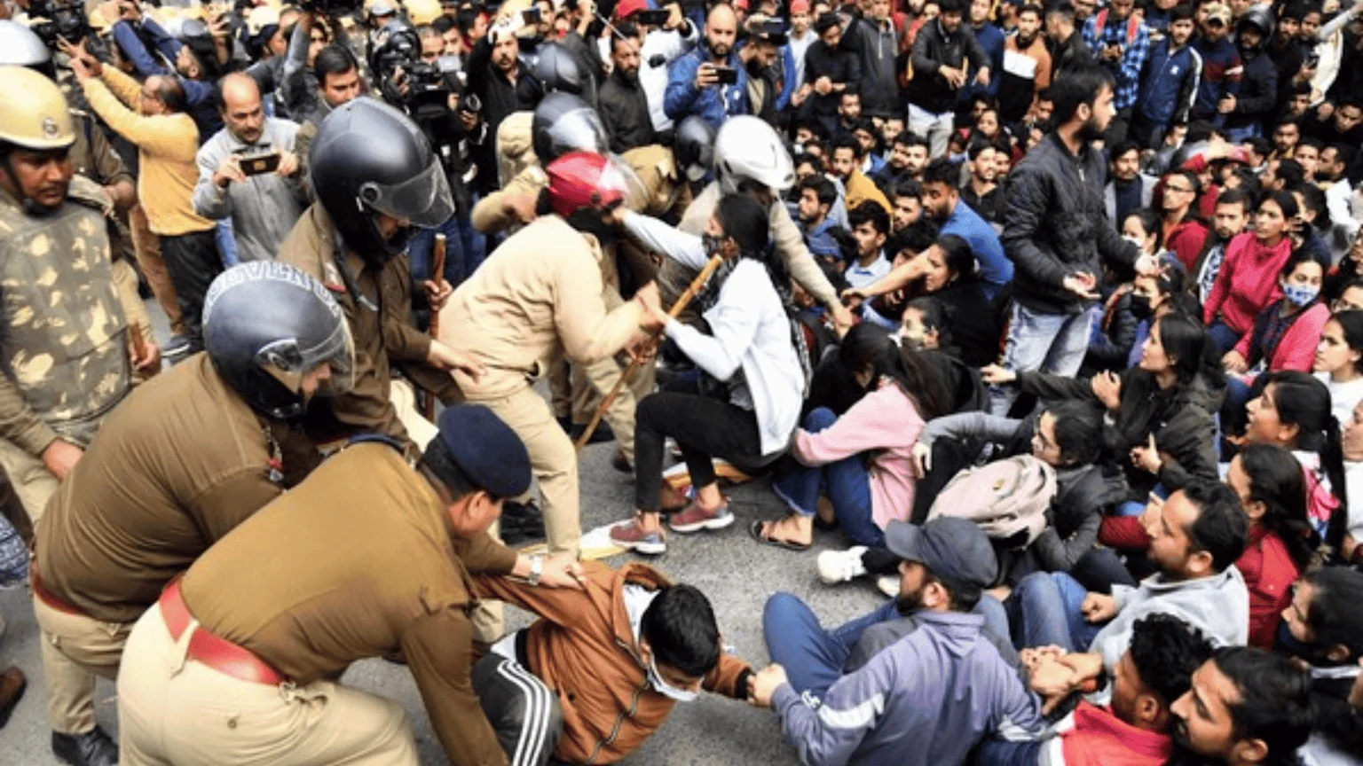 Police officers struggle with and pull at several young protestors, some of whom are seated and resisting, amidst a large crowd.